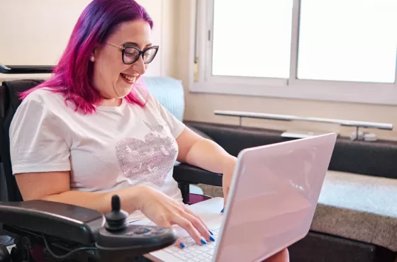 A woman with glasses and purple hair sitting in a wheelchair with a laptop in her lap. There is a bed and a window in the background.
