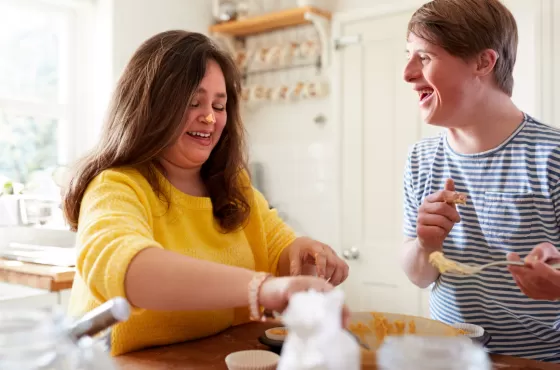 A woman with brown hair and a yellow jumper supporting a man with disability. They are both working together to make a cake mixture in a kitchen.