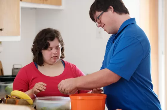 A woman with down syndrome in a red shirt talking to a man with down syndrome in a blue shirt, with a kitchen in the background.
