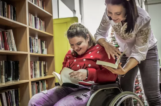 A woman in a wheelchair looking at a book in a library, with another woman looking over her shoulder.