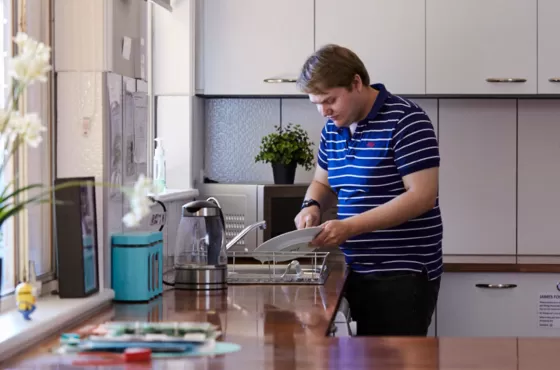 A man in a blue shirt and brown hair washing dishes with a kitchen in the background.