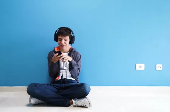 A young man with brown hair with headphones on listening to music on his phone. He is sitting on the ground with a blue wall behind him.