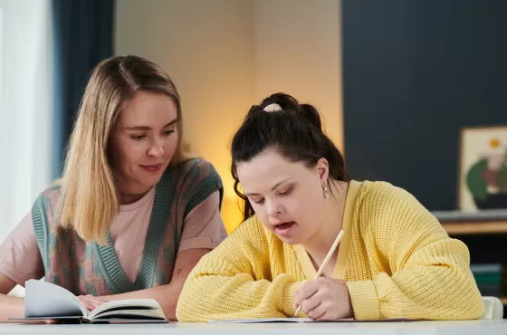 A woman with blonde hair sitting a table talking to a woman with down syndrome with a yellow cardigan. Both women are looking at books.