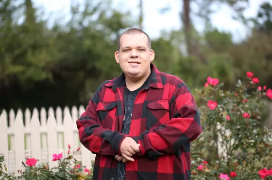 Man with shaved head wearing a red chequered jacket standing in a garden with a white picket fence in the background.