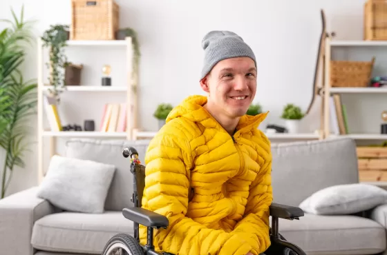 A man in a yellow jacket in a wheelchair with a sofa and bookcase in the background.