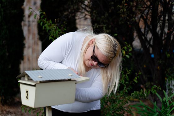 A young woman with blonde hair wearing a white shirt and black pants at a letterbox looking at an envelope.